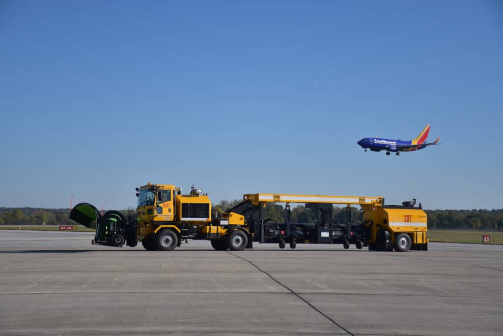 A large snow plow machine with areas for a broom to connect in the middle rides along the taxiway in Columbus, Ohio as a Southwest Airlines Boeing 737 comes in to land overhead.