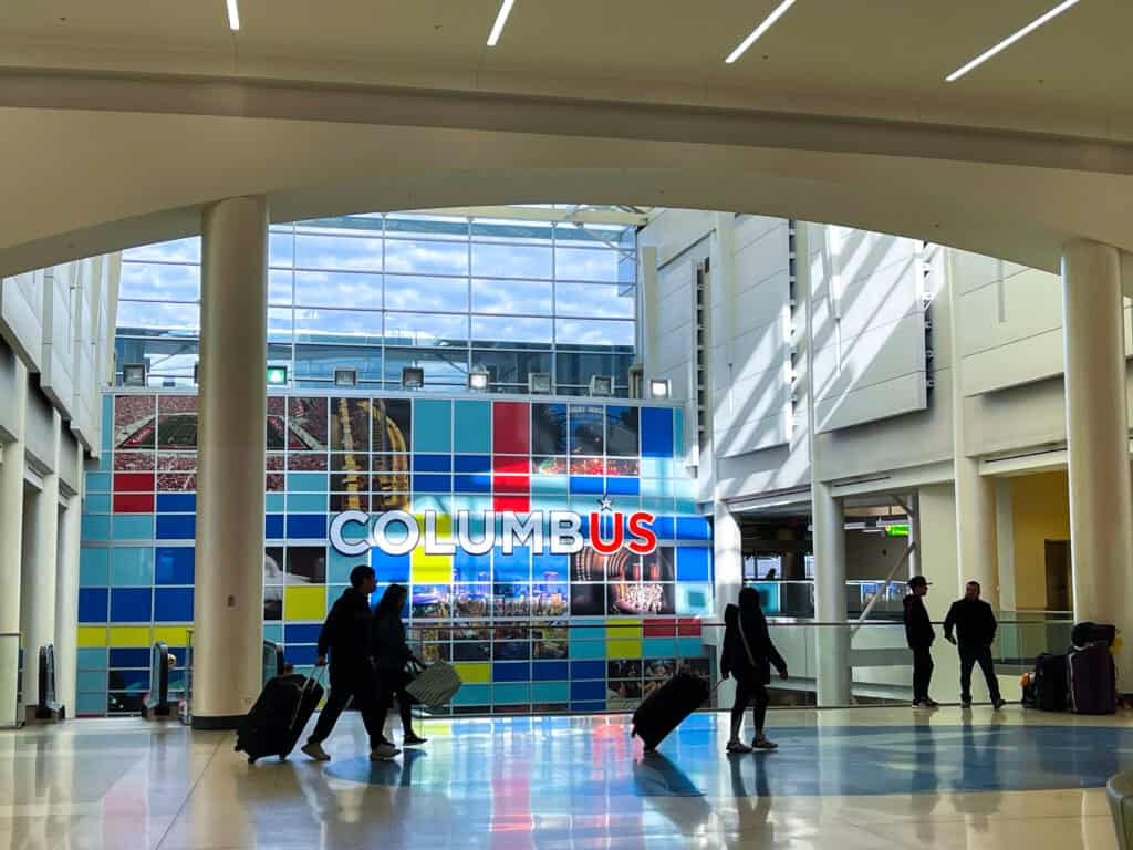 People walking in the ticket lobby at John Glenn Columbus International. Sun shines across a mosaic background.