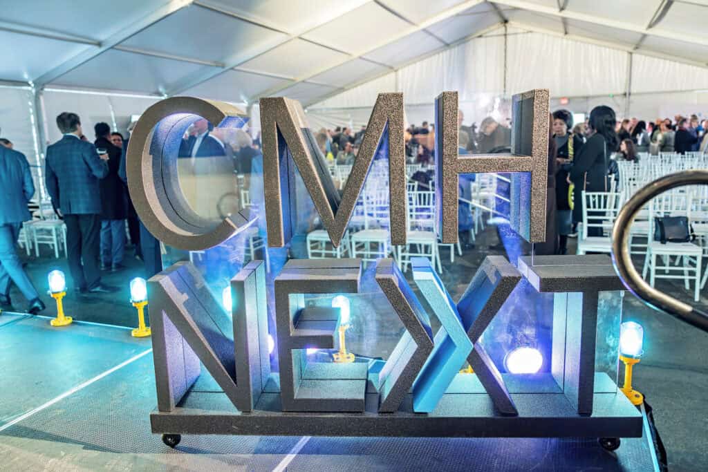 Decorative "CMH NEXT" sign on display at an outdoor event with people mingling in the background under a tent.