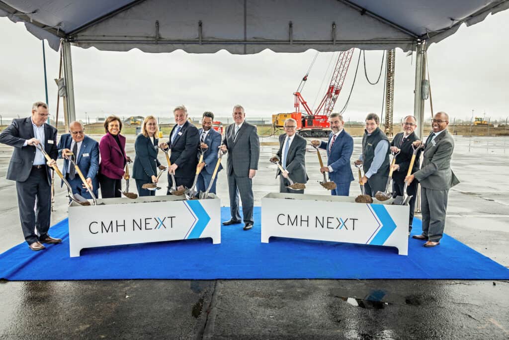 A group of eleven individuals wearing formal attire participating in a groundbreaking ceremony under a tent. They are holding shovels and posing for the photo with a large banner reading "CMH NEXT" in front of them. The setting includes an overcast sky and construction equipment in the background.