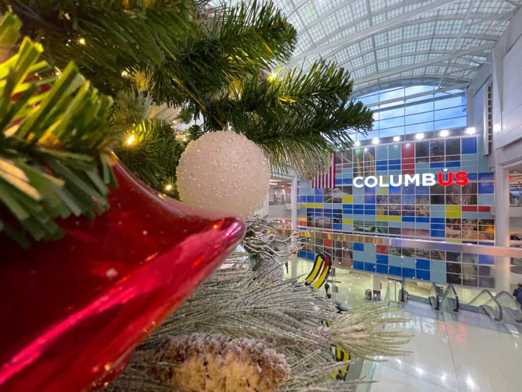 Close-up of a Christmas decoration with a red bell and white bauble on a tree, overlooking an indoor area with "COLUMBUS" signage in the background.