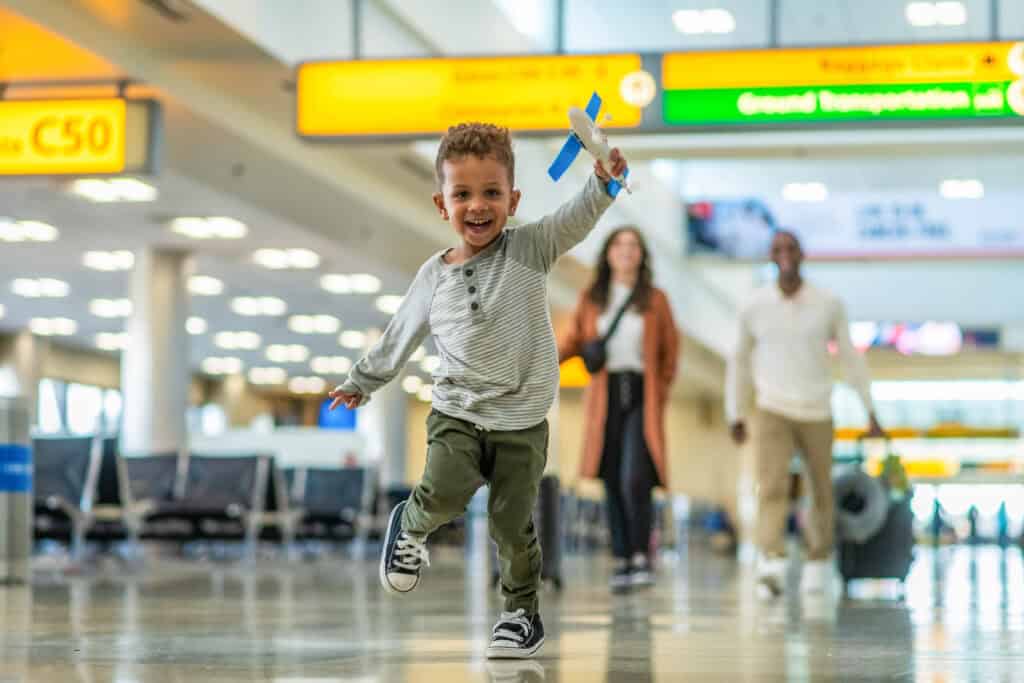 A little boy runs through the airport terminal, holding onto a small toy airplane. His parents are in the background, smiling while watching him as they walk along.