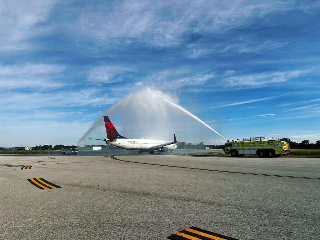 A Delta Air Lines aircraft taxis through two plumes of water being sprayed from fire trucks before takeoff.