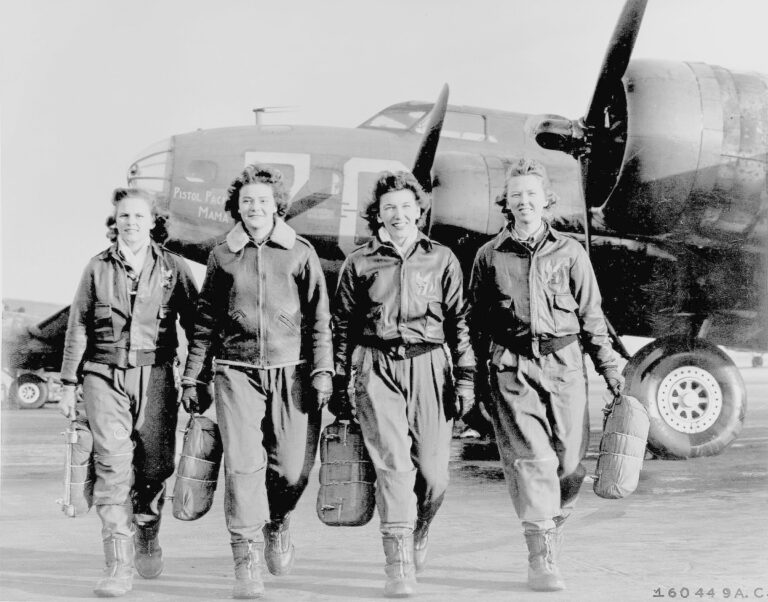 Four members of the Women Airforce Service Pilots (WASP) walking together in front of a military aircraft, each carrying a helmet and smiling, with "Pistol Packin' Mama" painted on the airplane's nose.