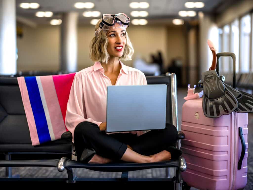 Person sits cross-legged on an airport bench, holding a laptop, with a pink suitcase and snorkeling gear beside them.