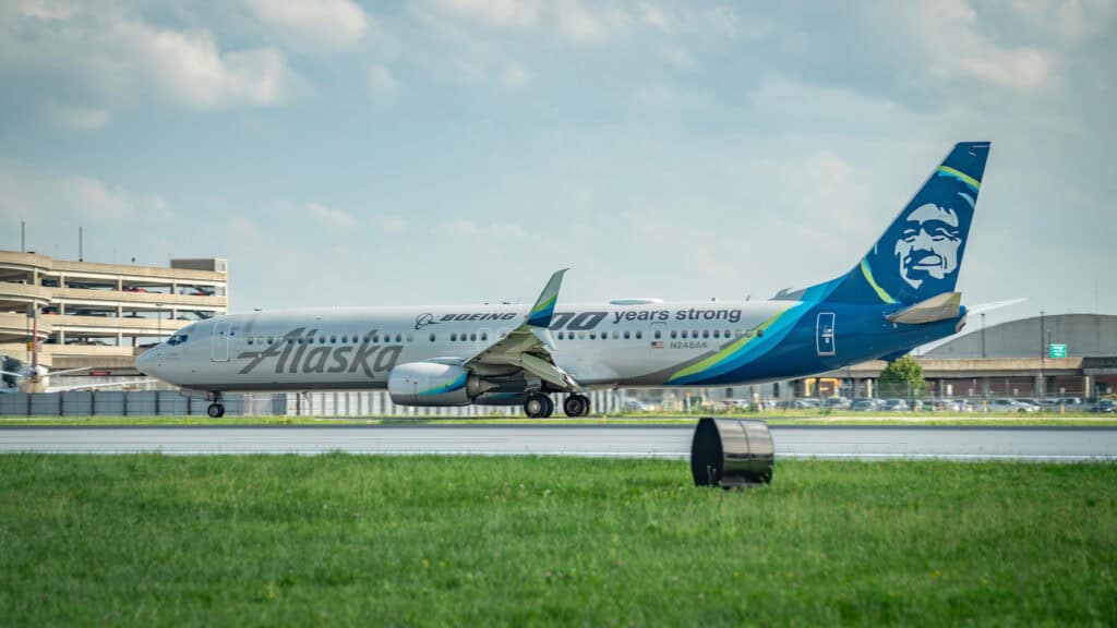 An Alaska Airlines airplane marked with "100 years strong" on the fuselage, parked on a runway with grass and an airport structure in the background.