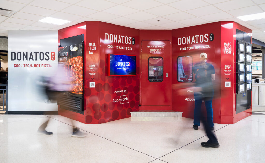 A bright red Donatos automated pizza kiosk inside John Glenn Columbus International Airport. The cube-shaped station displays signage reading “Cool Tech. Hot Pizza.” with digital screens, ordering instructions, and windows showing the robotic pizza-making equipment inside. Several blurred travelers walk past the kiosk, while one person stands near the ordering area.