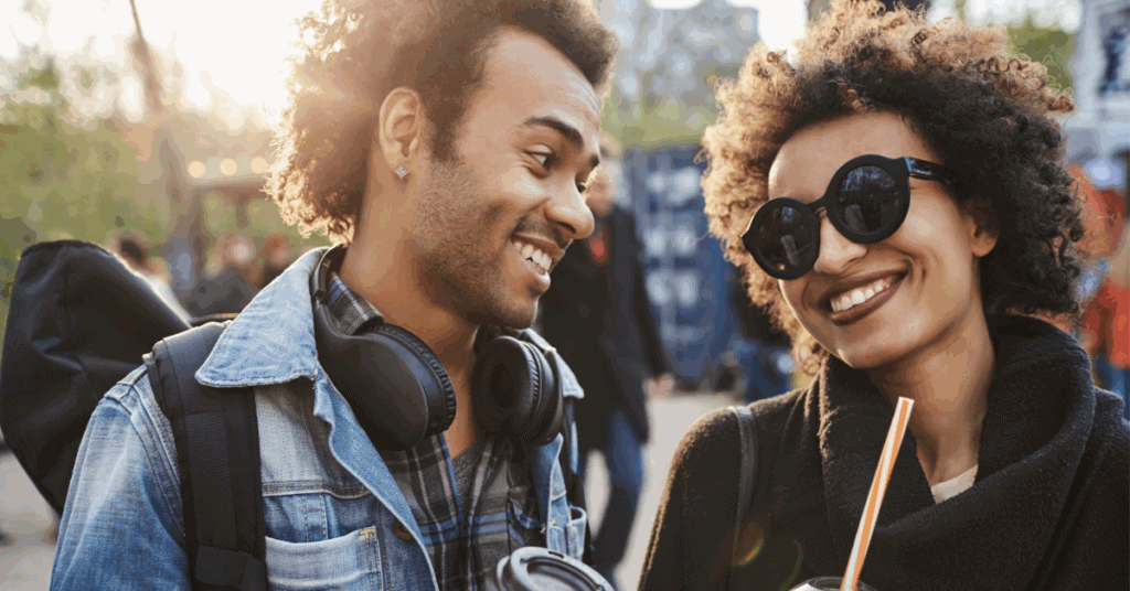 Two people enjoying a sunny day outdoors, smiling and walking together with one person wearing sunglasses and headphones around their neck.