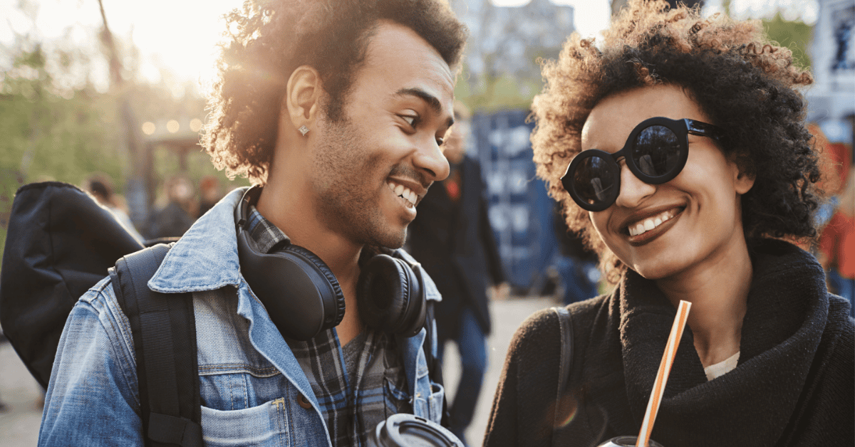Two people enjoying a sunny day outdoors, smiling and walking together with one person wearing sunglasses and headphones around their neck.