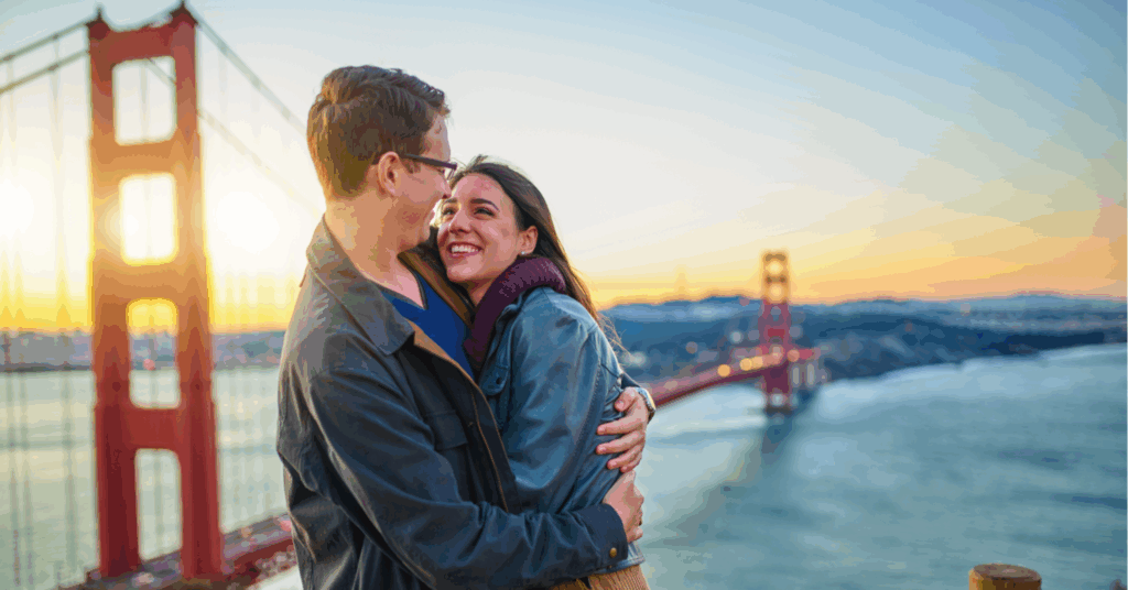 Two people hugging and smiling in front of the Golden Gate Bridge at sunset.