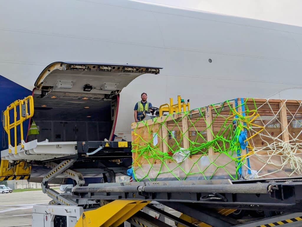 Ground crew member operates an electric cargo loader to move a wooden crate toward an aircraft’s cargo hold. The plane’s side cargo door is open with equipment extended.