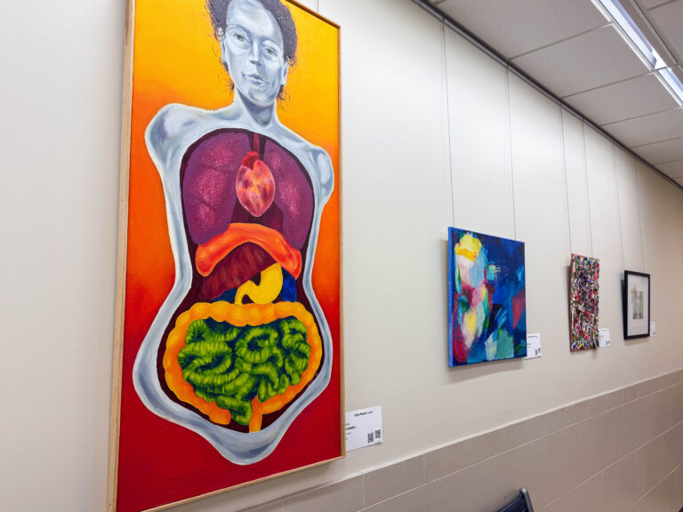 A close-up view of paintings in the Gateway to the Arts gallery, including a large piece depicting a human figure with internal organs in bright red, orange, and green tones. Other abstract and mixed-media works line the wall.
