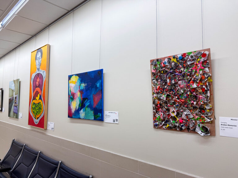 A row of colorful paintings displayed along a hallway at John Glenn Columbus International Airport’s Gateway to the Arts gallery. The works range from abstract and figurative pieces to a mixed-media mosaic filled with vibrant textures and shapes.