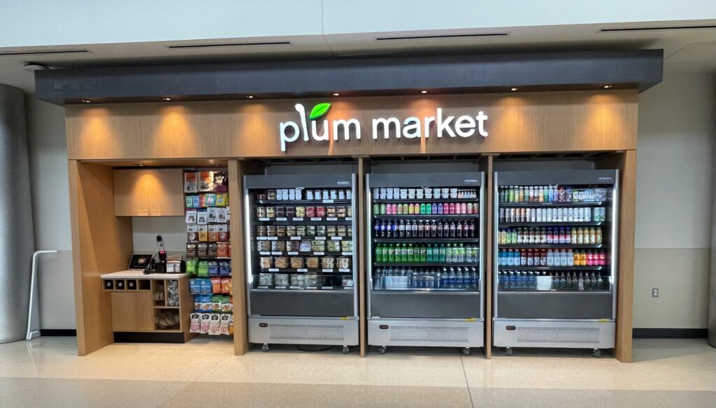 A Plum Market kiosk with refrigerated food items and drinks, along with snacks on the wall next to a cash register.