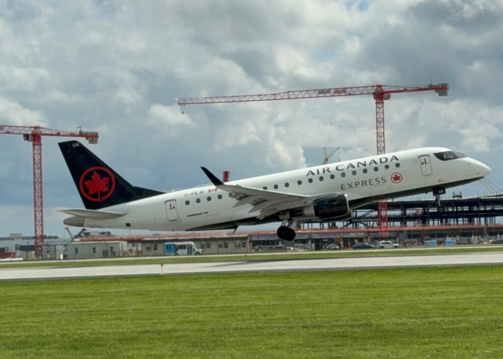 Air Canada Express Embraco 175 aircraft (registration C-FEJF) lifting off from a runway, with red construction cranes and a partially-built terminal structure visible in the background under a partly cloudy sky.