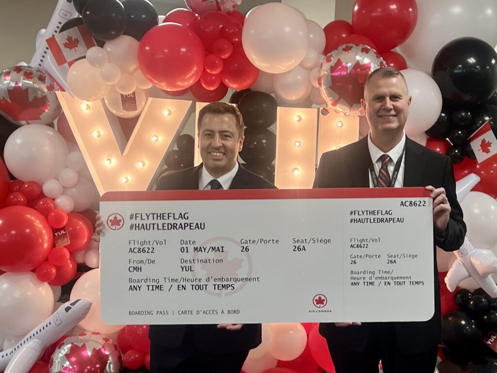 Two men in dark business suits smiling and holding a large oversized Air Canada boarding pass for flight AC8622 from CMH to YUL on 01 May, featuring the hashtags #FLYTHEFLAG and #HAUTLEDRAPEAU. They stand in front of an illuminated marquee "YUL" sign surrounded by a balloon arch in red, white, and black, with Canadian flag accents and inflatable airplane decorations.