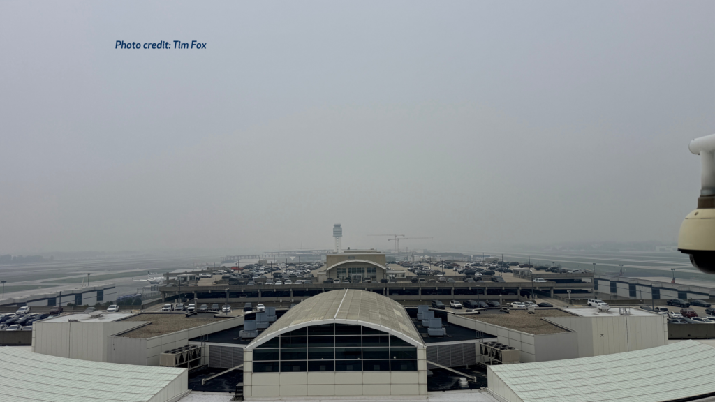 View of John Glenn Columbus International Airport from an elevated vantage point on a hazy day, with wildfire smoke reducing visibility across the airfield. The terminal roof, parking garage, control tower, runways, and construction cranes are visible beneath a gray sky. Photo credit: Tim Fox.