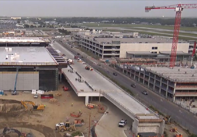 Aerial view of CMH Next construction at John Glenn Columbus International Airport, showing the elevated roadway, new terminal and parking structures, construction cranes, equipment, and surrounding airport grounds.