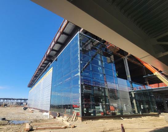 Close-up view of the CMH Next construction site, showing newly installed glass exterior panels on part of the new terminal, with exposed steel framing and ongoing construction visible around the building.