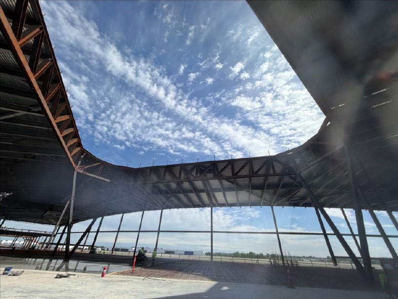 Interior view of the new CMH terminal under construction, looking up at the sweeping steel roof structure and large open window framing with blue sky visible overhead.