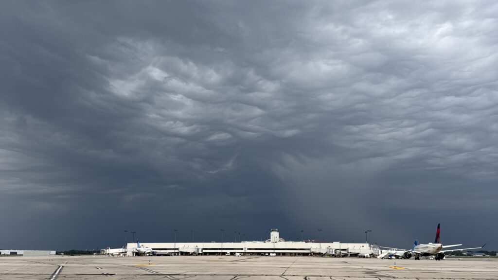 Wide view of an airport airfield and terminal beneath dark, dramatic storm clouds. Several commercial aircraft are parked at gates in the distance as heavy gray clouds fill most of the sky, signaling approaching severe weather.