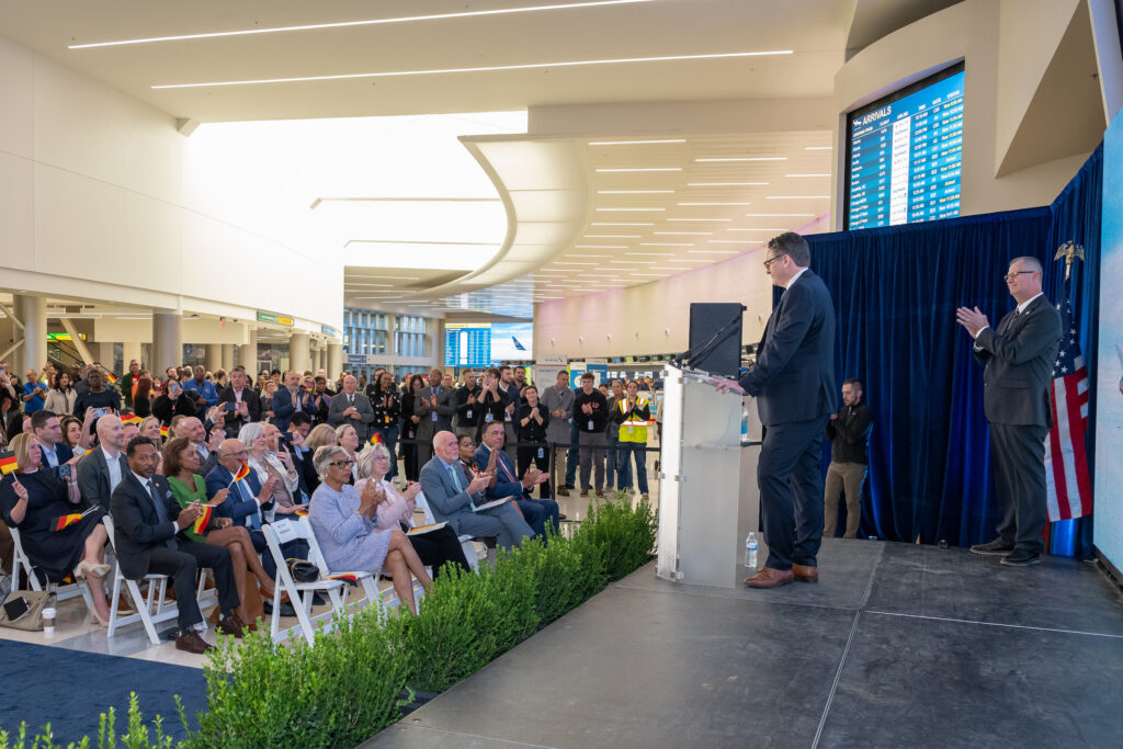 Airport employees stand along the perimeter of the seated event area, listening as Lufthansa’s Derk Janzen speaks onstage.