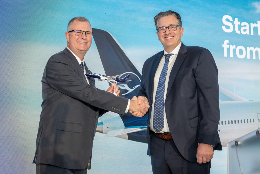 Two smiling men in suits shake hands, with one holding a model Lufthansa airplane, in front of a Lufthansa aircraft backdrop at the Columbus–Frankfurt service announcement.