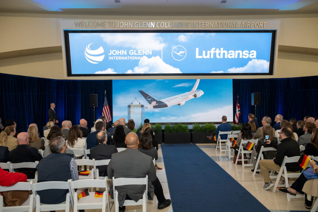 Columbus Regional Airport Authority CEO Daren Griffin speaks from a podium onstage as a video of Lufthansa’s Boeing 787-9 plays on a large screen behind him. A seated crowd watches the presentation.