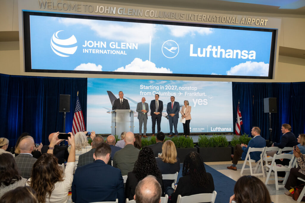 Attendees watch a speaker at a podium and four people onstage at John Glenn Columbus International Airport. Screens display the airport and Lufthansa logos and announce nonstop Columbus–Frankfurt service starting in 2027.