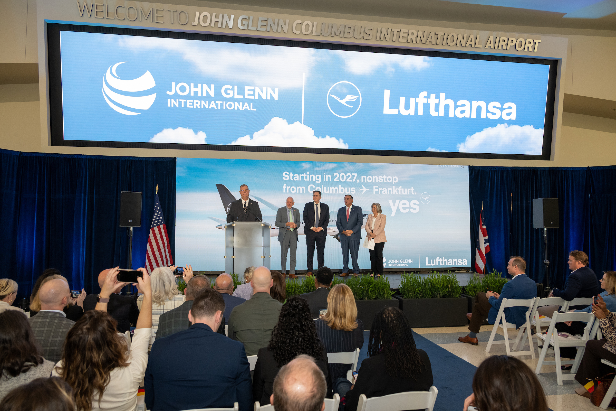 Attendees watch a speaker at a podium and four people onstage at John Glenn Columbus International Airport. Screens display the airport and Lufthansa logos and announce nonstop Columbus–Frankfurt service starting in 2027.