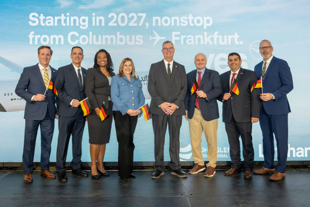 Columbus Regional Airport Authority’s executive team stands onstage holding German flags. From left: John McClelland, Chief Communications and Public Affairs Officer; Marc Sethan, Chief Operations Officer; Onnie Jacque, Legal Counsel; Cammi Wing, Chief People Officer; Daren Griffin, CEO; Tom McCarthy, Chief Planning & Engineering Officer; Fabio Spino, Chief Financial Officer; and Casey Denny, Chief Development Officer.